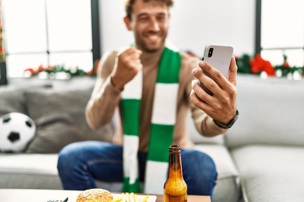 Foto grátis jovem hispânico apoiando partida de futebol usando smartphone sentado na decoração de natal em casa