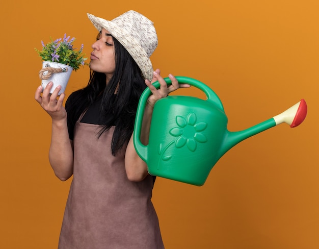 Foto grátis jovem jardineira caucasiana de uniforme e chapéu, segurando um vaso de flores e regador, cheirando flores