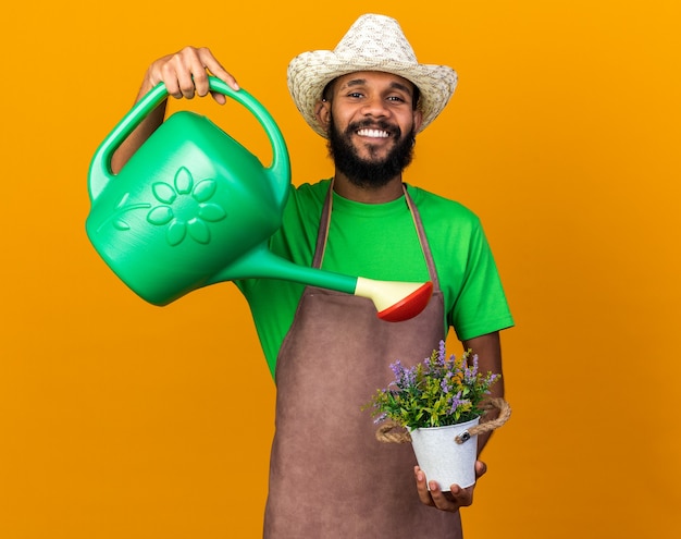 Foto grátis jovem jardineiro afro-americano sorridente, usando um chapéu de jardinagem, regando uma flor em um vaso de flores com um regador