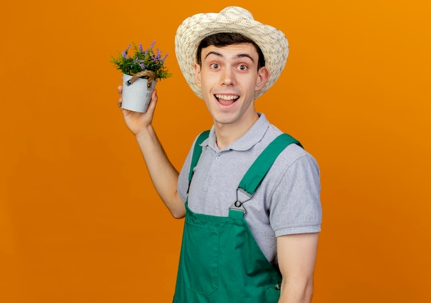 Foto grátis jovem jardineiro alegre usando chapéu de jardinagem segurando flores em um vaso de flores