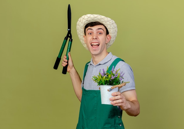 Foto grátis jovem jardineiro alegre usando chapéu de jardinagem segurando uma tesoura e um vaso de flores