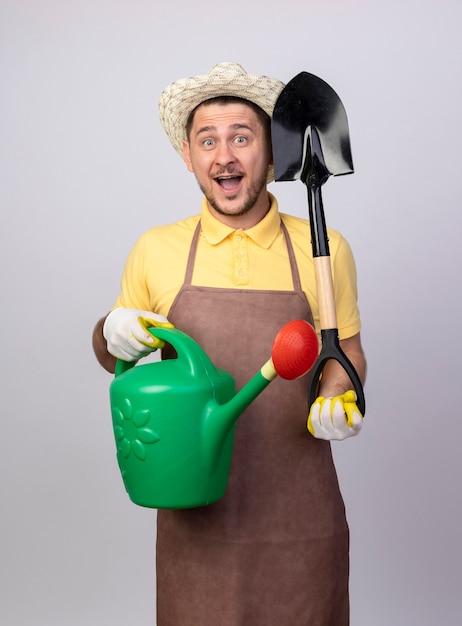 Foto grátis jovem jardineiro alegre vestindo macacão e chapéu em luvas de trabalho segurando um regador e uma pá, sorrindo com uma cara feliz