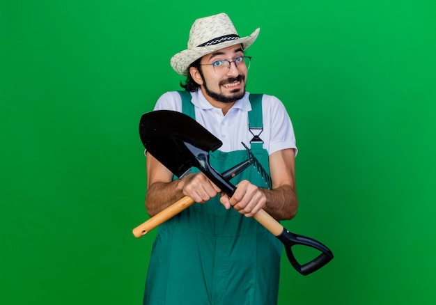 Foto grátis jovem jardineiro barbudo usando macacão e chapéu segurando um mini ancinho e uma pá confuso