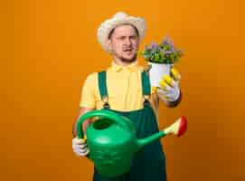 Foto grátis jovem jardineiro de macacão e chapéu segurando um regador, olhando para um vaso de planta com as duas mãos, ficando descontente e confuso em pé sobre a parede laranja