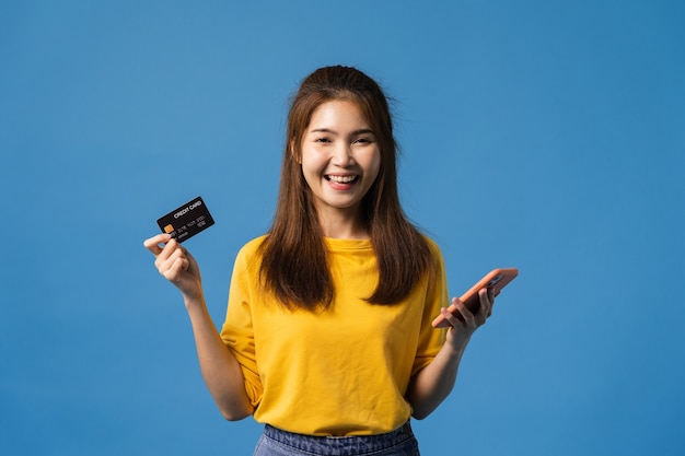 Foto grátis jovem senhora asiática usando telefone celular e cartão de banco de crédito com expressão positiva, vestida com um pano casual e olhando para a câmera isolada sobre fundo azul. mulher feliz adorável feliz alegra sucesso.