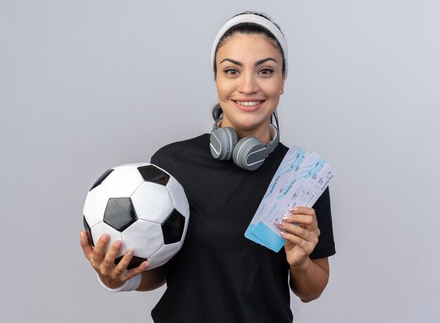 Jovem sorridente caucasiana esportiva usando bandana e pulseiras com fones de ouvido ao redor do pescoço segurando as passagens de avião e uma bola de futebol olhando para a frente, isolada na parede branca