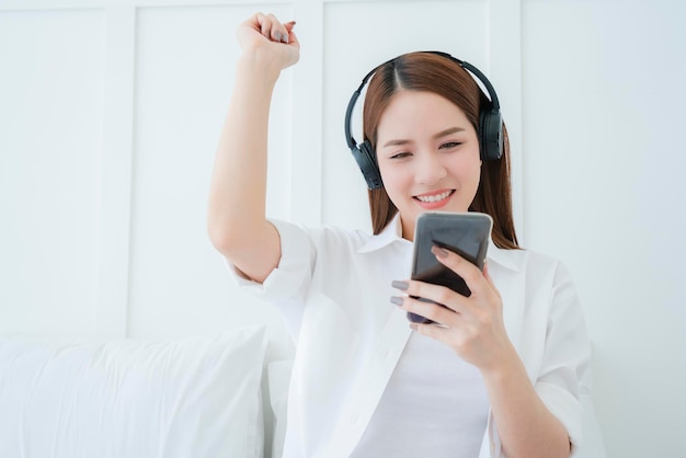 Foto grátis linda e atraente mulher asiática ouve música do fone de ouvido, segure a mão do smartphone, quarto branco retrato de fundo da mulher asiática de camisa branca de cabelos longos