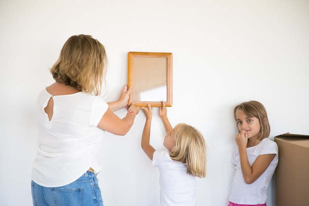 Linda garota colocando um quadro na parede branca com a ajuda da mãe