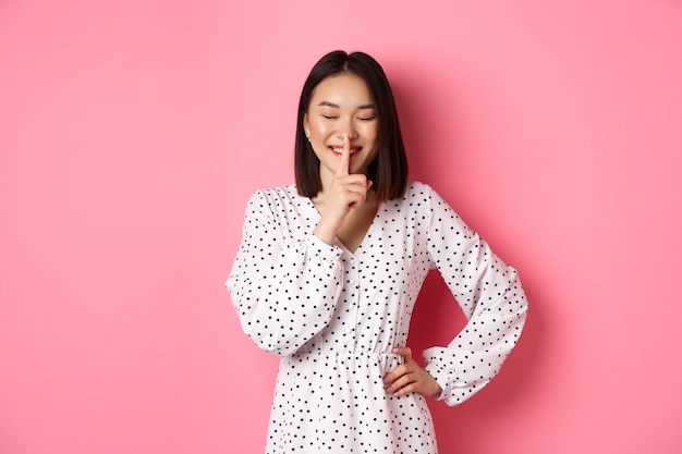 Foto grátis linda mulher coreana em um vestido da moda pedindo para guardar segredo com um sorriso suave e olhos fechados ...