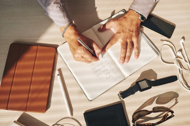 Mãos de homem escrevendo no diário e gadgets na mesa