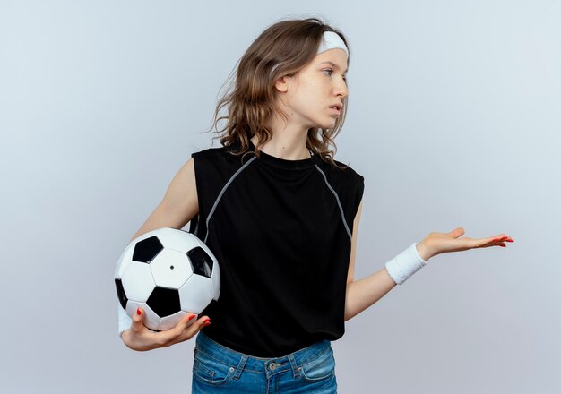 Menina jovem fitness em roupa esportiva preta com tiara segurando uma bola de futebol, olhando para o lado com o braço estendido enquanto perguntando em pé sobre uma parede branca