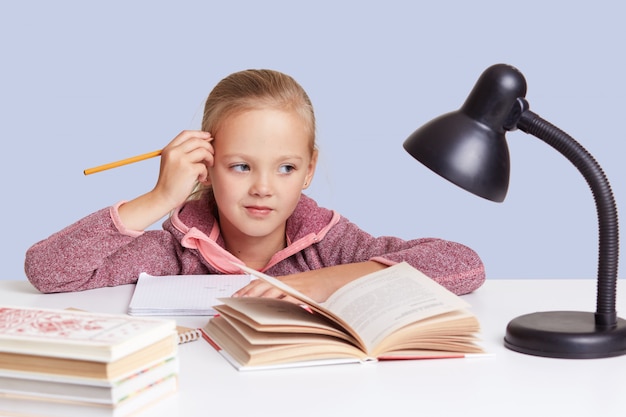 Foto grátis menininha mantém a mão perto da cabeça, olha com expressão pensativa, pensa na tarefa de casa, usa lâmpada de leitura. crianças, educação e conceito de escolaridade.