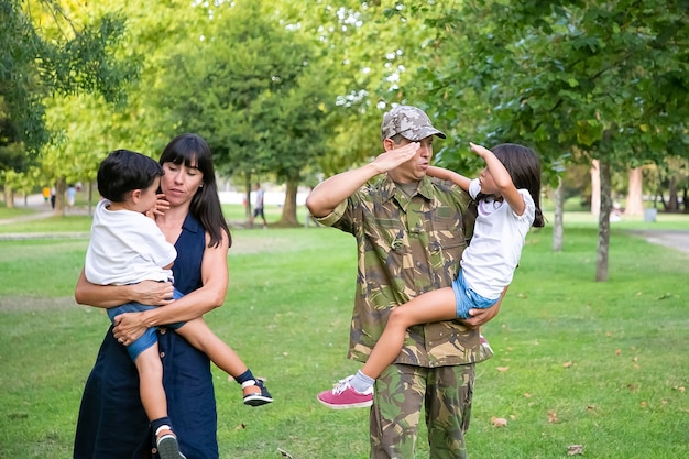 Foto grátis militar positivo andando no parque com sua esposa e filhos, ensinando a filha a fazer o gesto de saudação do exército. comprimento total, vista traseira. reunião de família ou conceito de pai militar