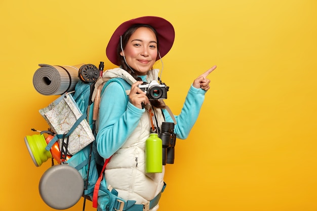 Foto grátis mochileira ativa aponta o dedo indicador para o espaço da cópia de lado, segura uma câmera retro, tira fotos, carrega mochila, binóculos e garrafa térmica, usa roupas casuais