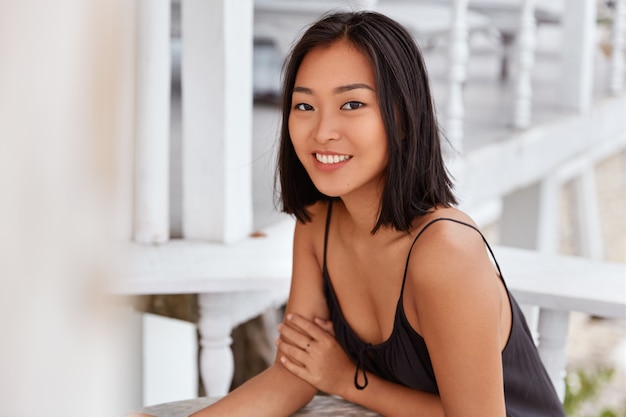 Foto grátis mulher asiática satisfeita com um sorriso largo, penteado curto, vestido casualmente, senta-se à mesa do café, gosta do tempo de lazer. linda mulher japonesa descansando sozinha em restaurante