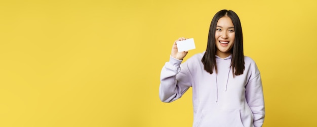 Foto grátis mulher asiática sorridente de compras mostrando um cartão na mão em pé sobre um fundo amarelo