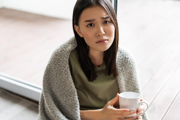 Foto grátis mulher asiática triste e sombria sentada em casa perto da janela segurando uma xícara de café e franzindo a testa ...