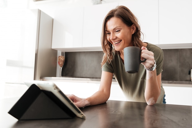 Mulher casual feliz tomando café e usando computador tablet na cozinha