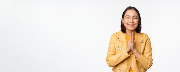 Foto grátis mulher coreana feliz olhando esperançosa pedindo ajuda favor implorando de pé com gesto de namaste e sorrindo em pé sobre fundo branco