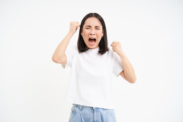 Foto grátis mulher coreana furiosa gritando e gritando, parecendo magoada e frustrada apertando as mãos em pé sobre wh