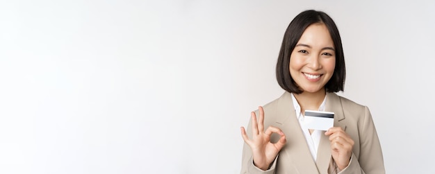 Foto grátis mulher corporativa asiática sorridente de funcionário de escritório mostrando cartão de crédito e sinal de ok recomendando banco em pé sobre fundo branco em terno bege