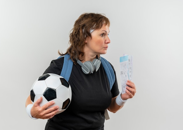 Mulher de meia-idade, esportiva, com uma camiseta preta com bandana e mochila segurando passagens aéreas e uma bola de futebol olhando para o lado com expressão pensativa no rosto em pé sobre uma parede branca