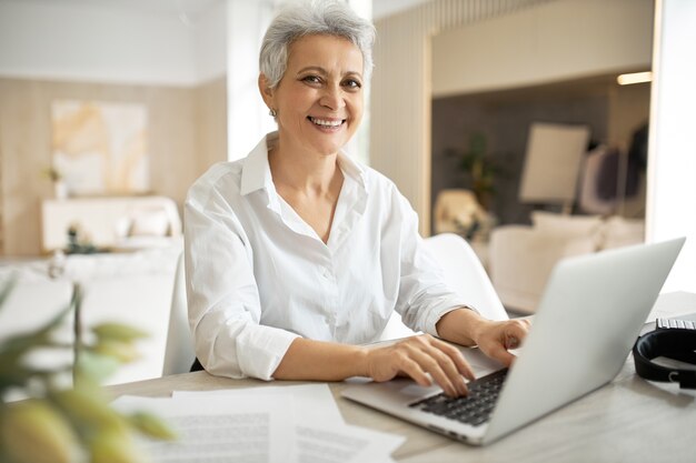 Mulher de negócios madura elegante com corte de cabelo curto sentada em frente ao laptop, olhando para a tela com a boca aberta como se estivesse dizendo algo