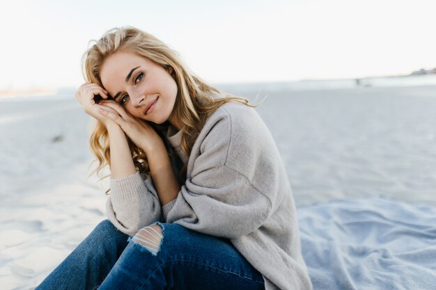 Mulher de olhos verdes em jeans e suéter está sentada na areia, com os joelhos apoiados no mar.