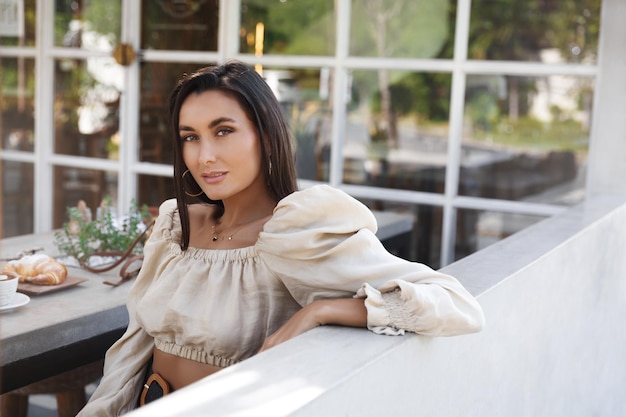 Foto grátis mulher elegante atraente senta-se à mesa de café ao ar livre comendo croissant e bebendo café modelo feminino sexy olha câmera inclinada na cerca enquanto desfruta de refeição no restaurante de café