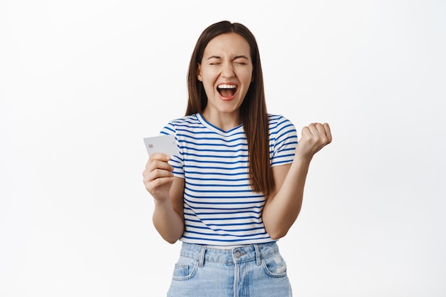 Foto grátis mulher emocional grita de felicidade e satisfação, mostrando segurando o cartão de crédito, cerrando os punhos como campeã, comemorando, em pé sobre uma parede branca