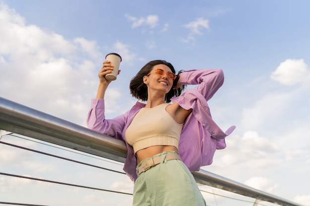 Foto grátis mulher feliz com a figura perfeita em uma elegante camisa roxa grande, desfrutando de uma xícara de café enquanto caminha na ponte moderna