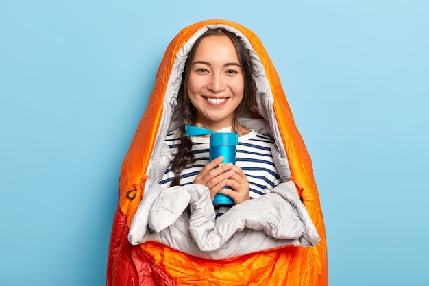 Foto grátis mulher feliz com longa trança, fica embrulhado em saco de dormir, segura a garrafa térmica com bebida quente, gosta de viajar e fazer caminhadas