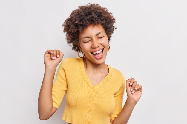 Foto grátis mulher feliz e relaxada pega um momento despreocupado desfruta da liberdade canta música mantém os braços levantados fecha os olhos dança ao som da música favorita usa camiseta amarela casual isolada na parede branca