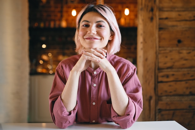 Foto grátis mulher jovem e alegre, positiva, com cabelo rosado, sentada em casa contra um fundo de luz dourada, com uma expressão facial feliz otimista, mantendo as mãos sob o queixo e sorrindo amplamente para a câmera