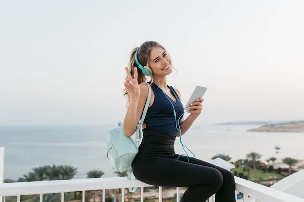 Foto grátis mulher jovem e atraente alegre no sportswear sorrindo, conversando no telefone, relaxando à beira-mar. nascer do sol pela manhã, modelo moderno, exercícios, música em fones de ouvido, clima alegre