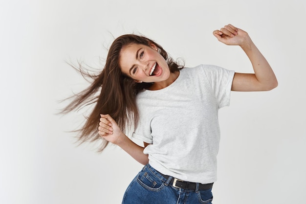 Foto grátis mulher jovem feliz dançando e chicoteando cabelos brilhantes e saudáveis, se divertindo, alegre em pé sobre uma parede branca