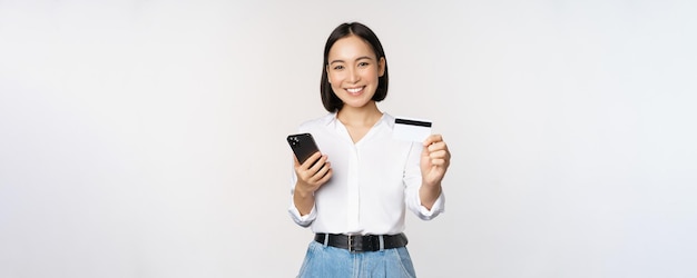 Foto grátis o conceito de compras on-line sorridente menina asiática moderna mostra que seu cartão de crédito possui pedido de telefone celular