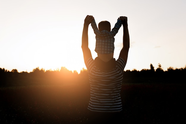 O pai e o filho que andam no campo no tempo do por do sol, menino que senta-se em equipa os ombros.