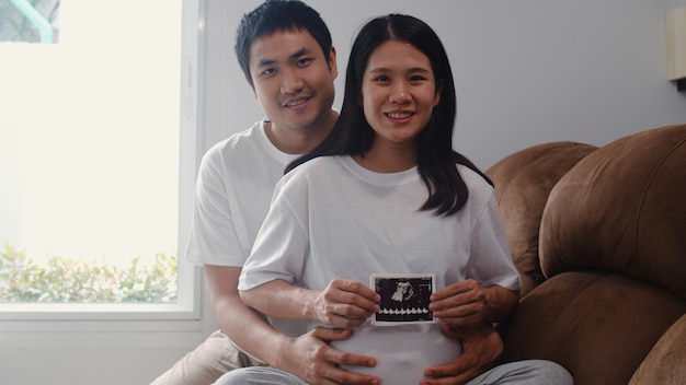 Os pares grávidos asiáticos novos mostram e olhando o bebê da foto do ultrassom na barriga. Mamãe e papai se sentindo feliz sorrindo pacífica enquanto cuida criança deitado no sofá na sala de estar em casa.