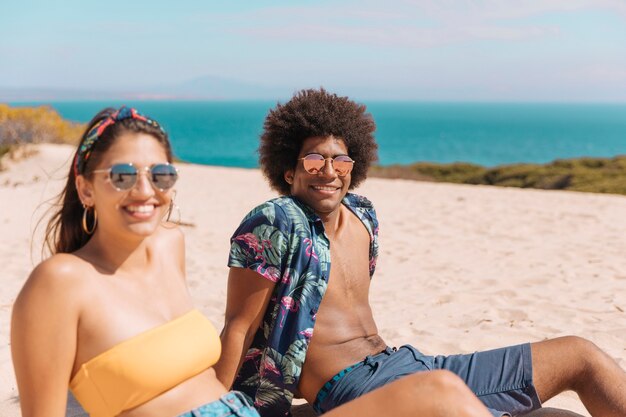 Par jovem, em, óculos de sol, sentando praia, sorrindo, e, olhando câmera