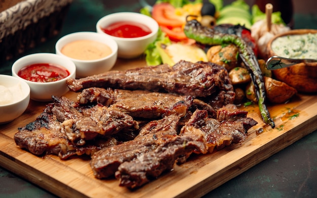 pedaços de bife de cordeiro com molhos, pimenta grelhada, salada fresca na placa de madeira