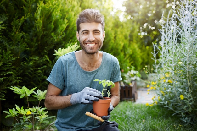 Foto grátis retrato ao ar livre de jovem homem barbudo bonito caucasiano em camisa azul e luvas, sorrindo na câmera, segurando o pote com flores nas mãos, trabalhando no jardim.