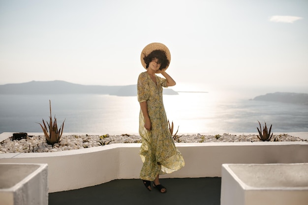 Foto grátis retrato completo da senhora morena feliz no vestido midi e velejador posando no terraço com vista para o mar mulher atraente no chapéu e roupa floral sorri no fundo do oceano