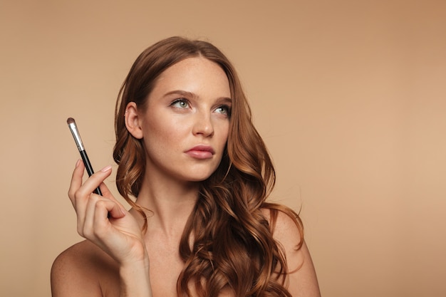 Foto grátis retrato da beleza da mulher pensativa de gengibre com cabelos longos, olhando para cima enquanto segura a escova de cosméticos