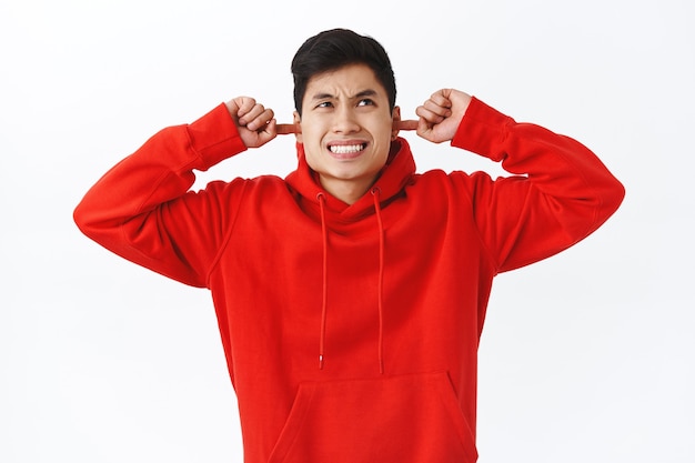 Foto grátis retrato da cintura de um jovem irritado, estudante asiático do sexo masculino, reclamando dos vizinhos barulhentos, ouvidos fechados e fazendo caretas, olhando para cima irritado, ouvir música alta irritante, parede branca.