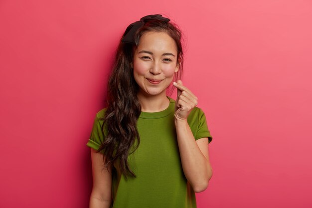 Retrato de adorável menina morena espalha amor e felicidade, mostra o sinal do coração, símbolo coreano de afeto com os dedos, usa uma camiseta verde, isolado sobre um fundo rosa brilhante