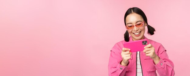 Foto grátis retrato de menina asiática feliz jogando no smartphone assistindo vídeos no aplicativo de celular sobre fundo rosa