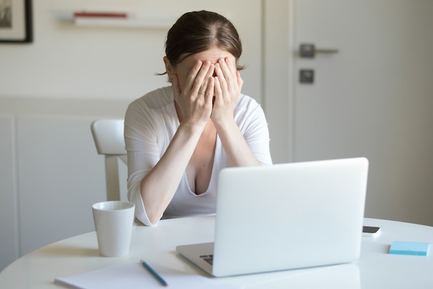 Retrato de mulher na mesa com laptop, mãos fechando rosto