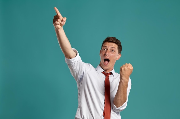 Foto grátis retrato de um homem moreno feliz com olhos castanhos, vestindo uma camisa branca clássica e gravata vermelha. ele está agindo como se tivesse ganhado algo posando em um estúdio contra um fundo azul. conceito de gesticulação