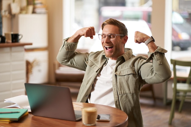 Foto grátis retrato de um jovem feliz com óculos alegrando-se sentado em um café assistindo a um jogo de esportes no laptop